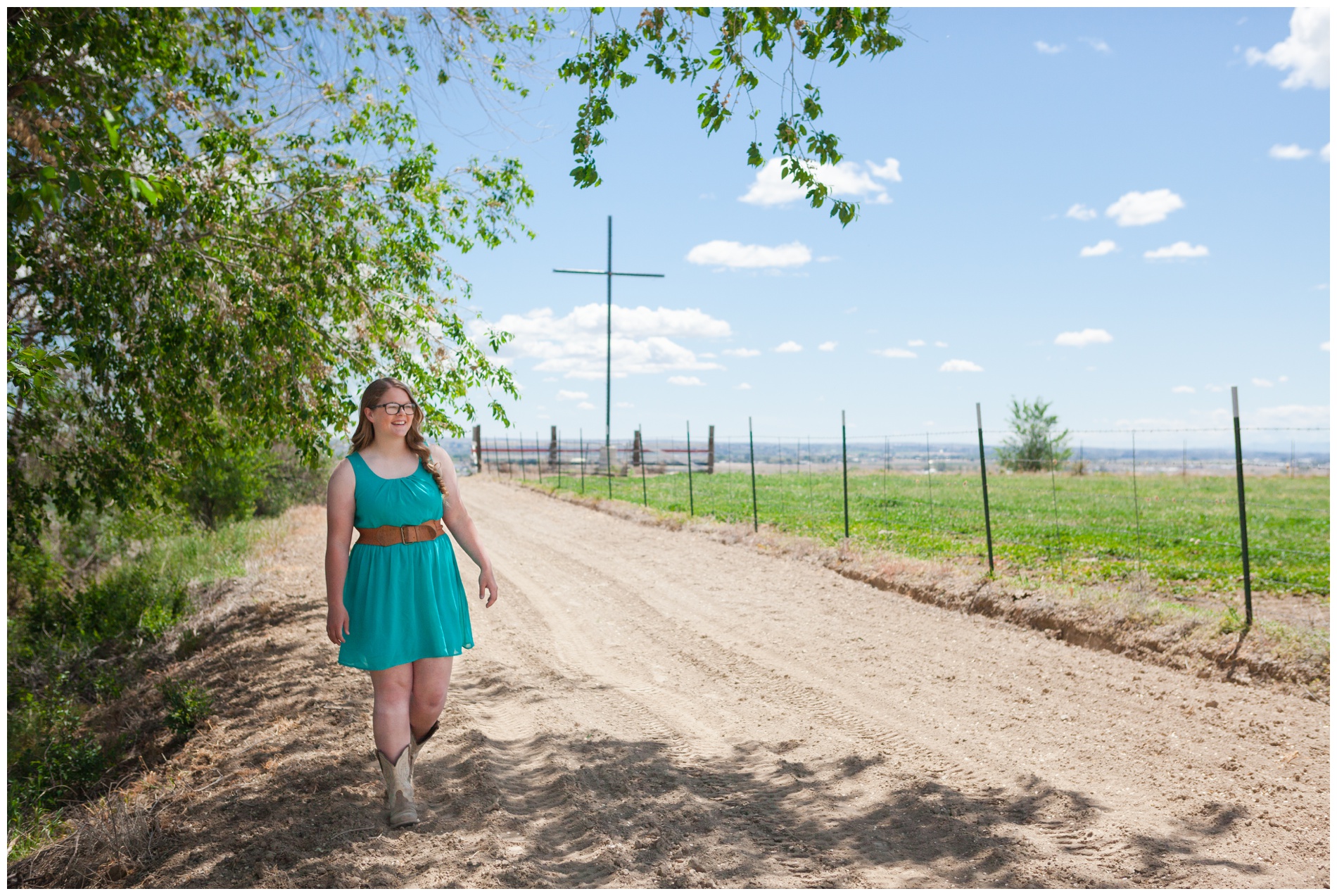 Picture of a high school senior girl walking down a dirt road with a large cross in the background.