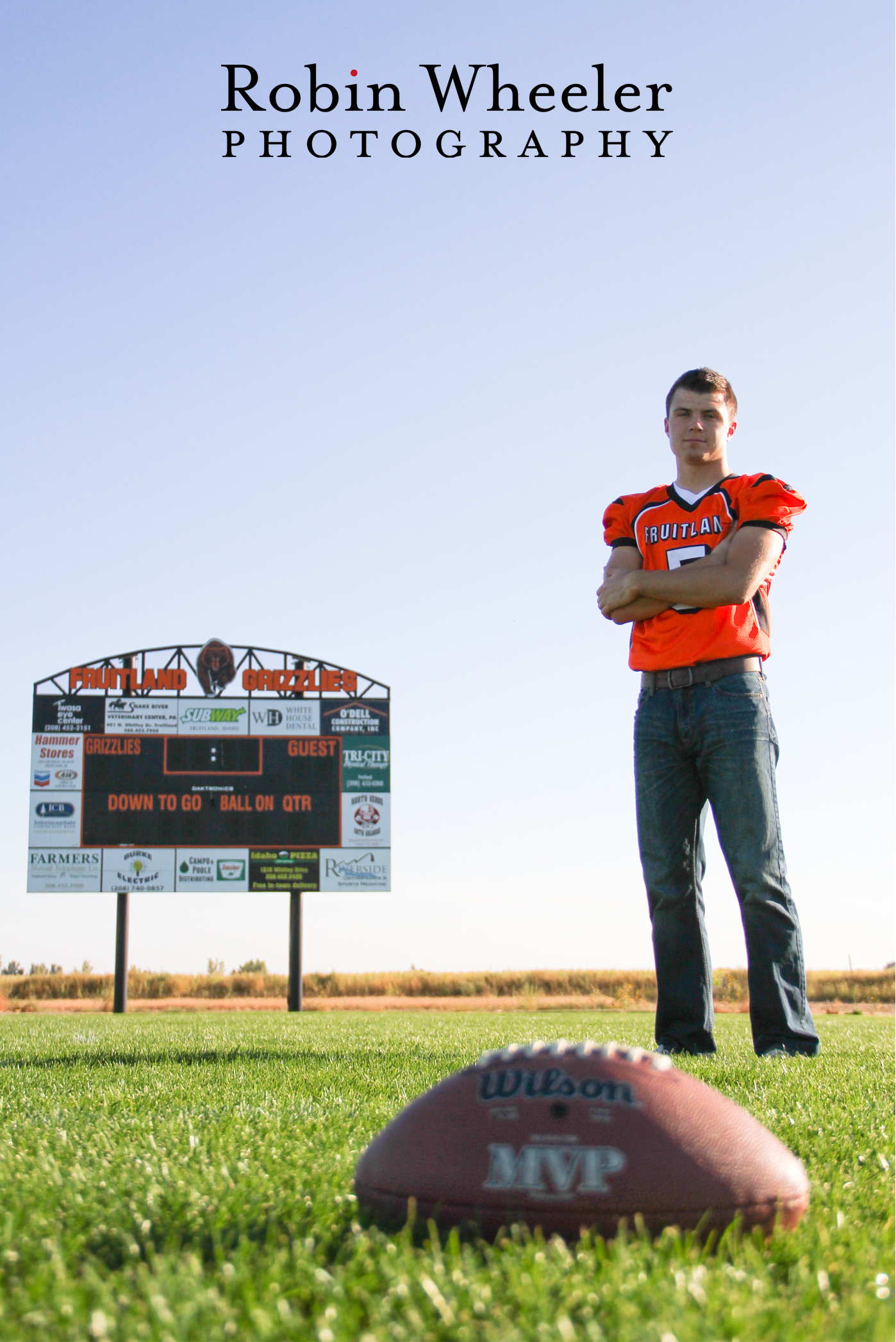 Senior football player standing in high school field with scoreboard in background and a football in the foreground.
