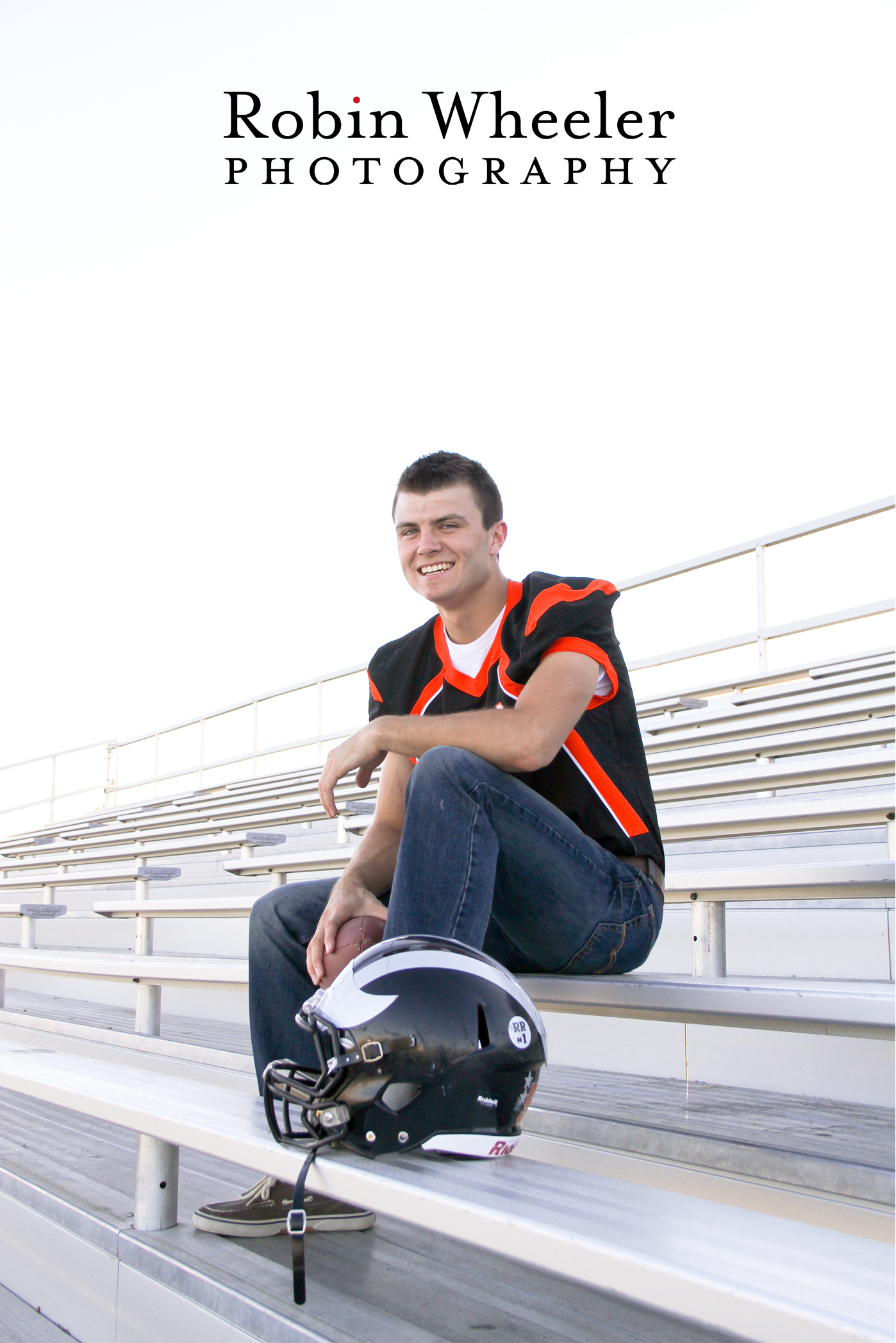 Photo of a high school senior in his football jersey, holding a football, sitting in the bleachers