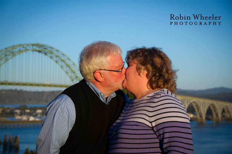 Family portrait of a couple kissing at Yaquina Bay State Recreation Site with the Yaquina Bay Bridge behind them, Newport, Oregon