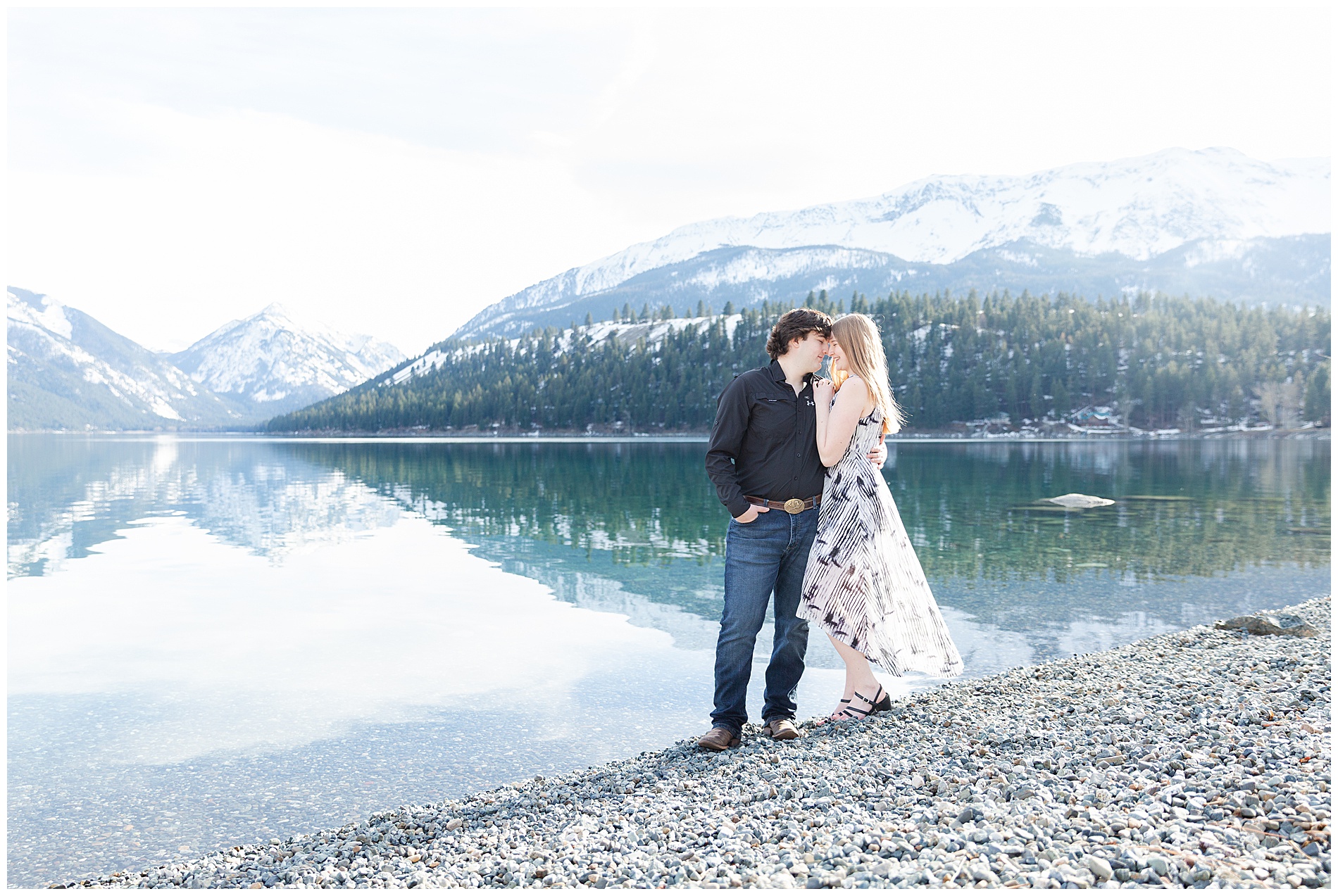 Epic shot of a couple standing on a rocky shore of Wallowa Lake with snow-capped Wallowa mountains behind