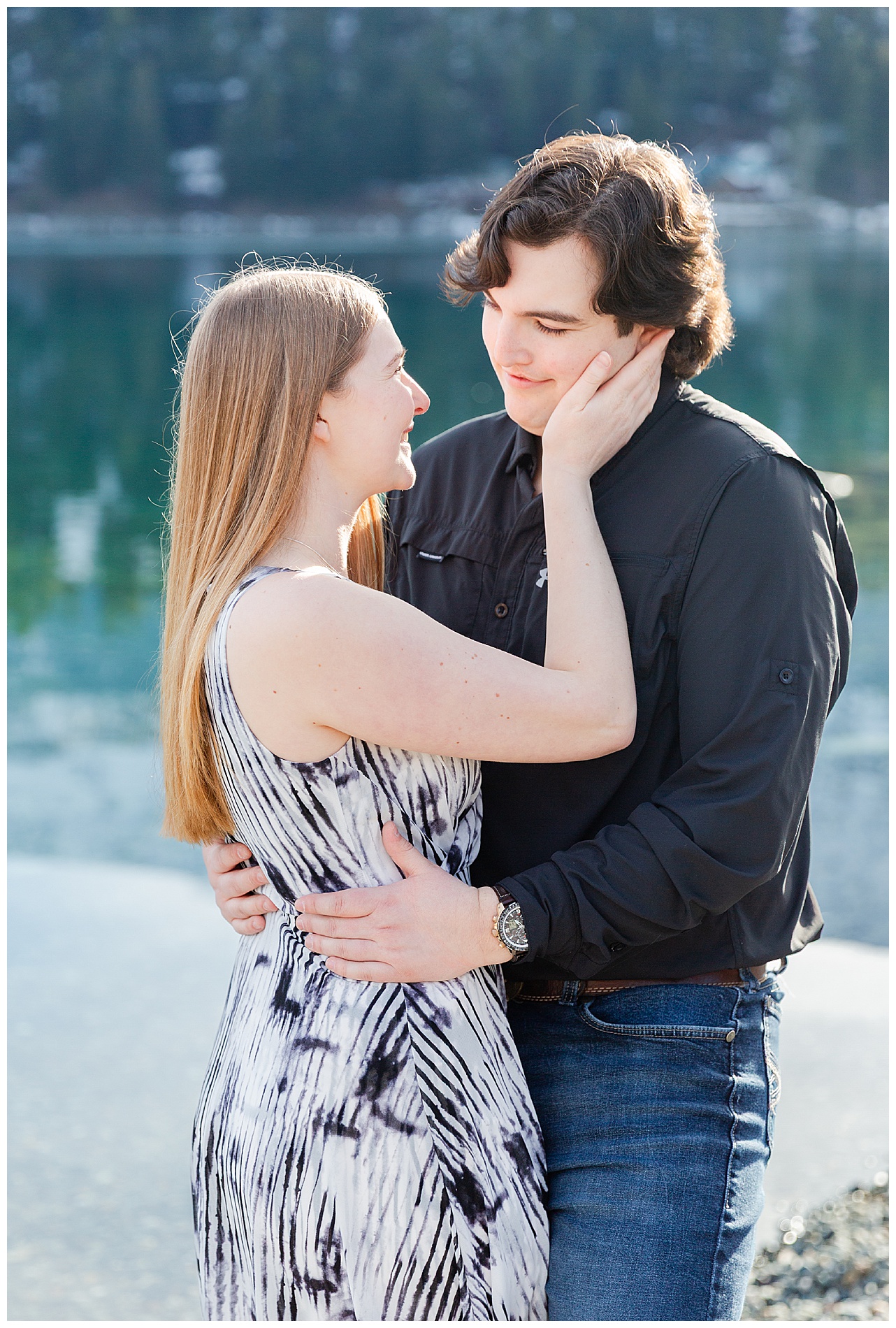 Engaged couple embracing with the mountains reflected in the water behind them