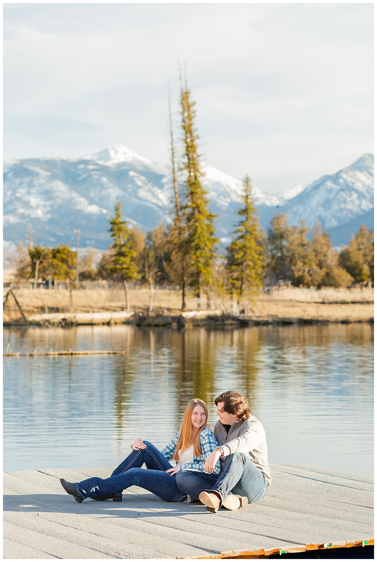The couple sits on a dock with evergreen trees and the Wallowa mountains behind them