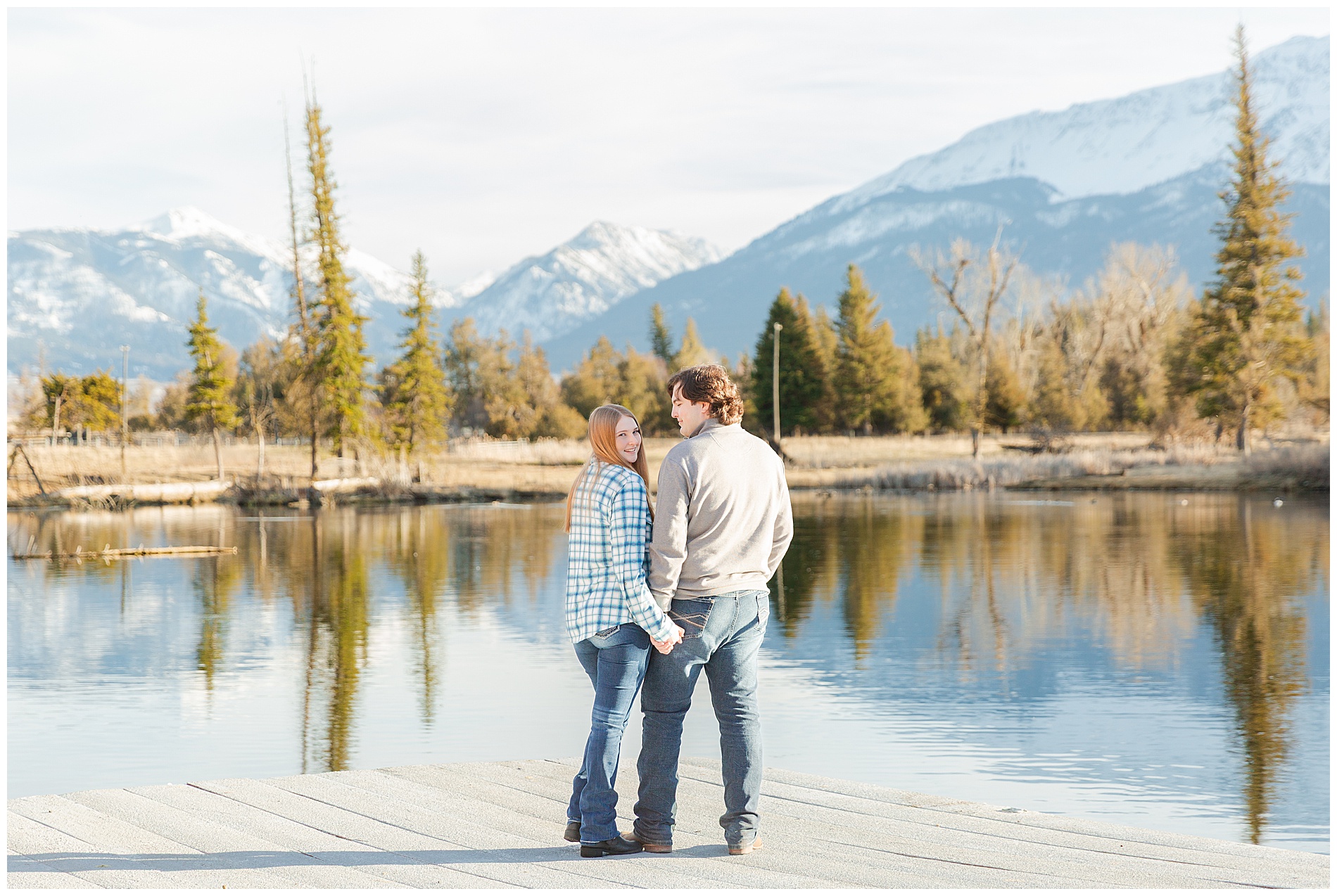 The couple standing on a dock with evergreen trees and the Wallowa mountains behind them
