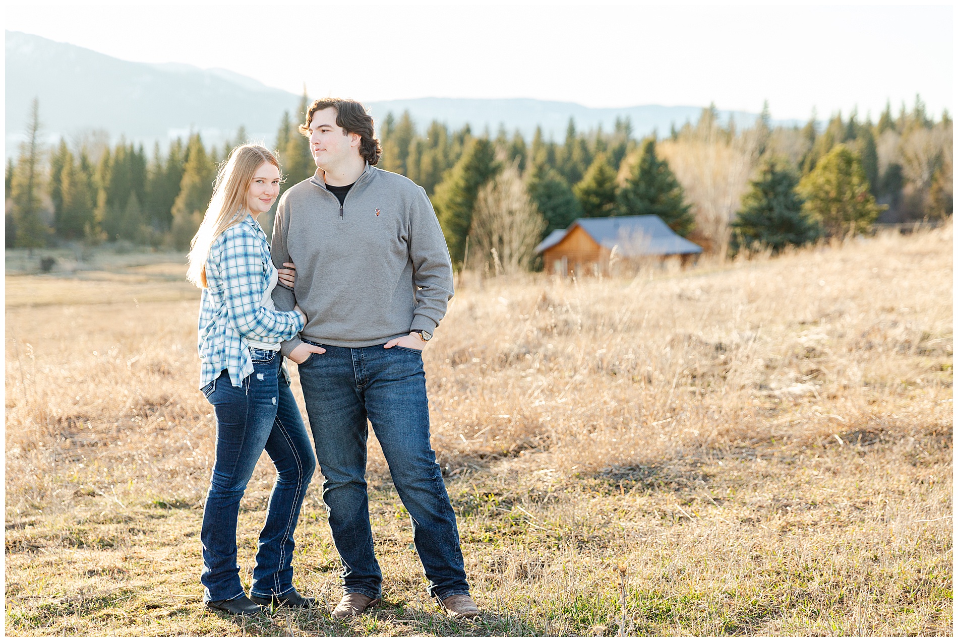 The couple stands together in a golden field with a trees and a cabin in the background