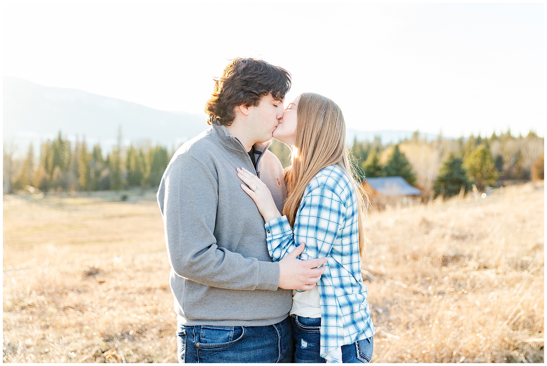 Engaged couple kissing with mountains, evergreens, and a cabin in the background