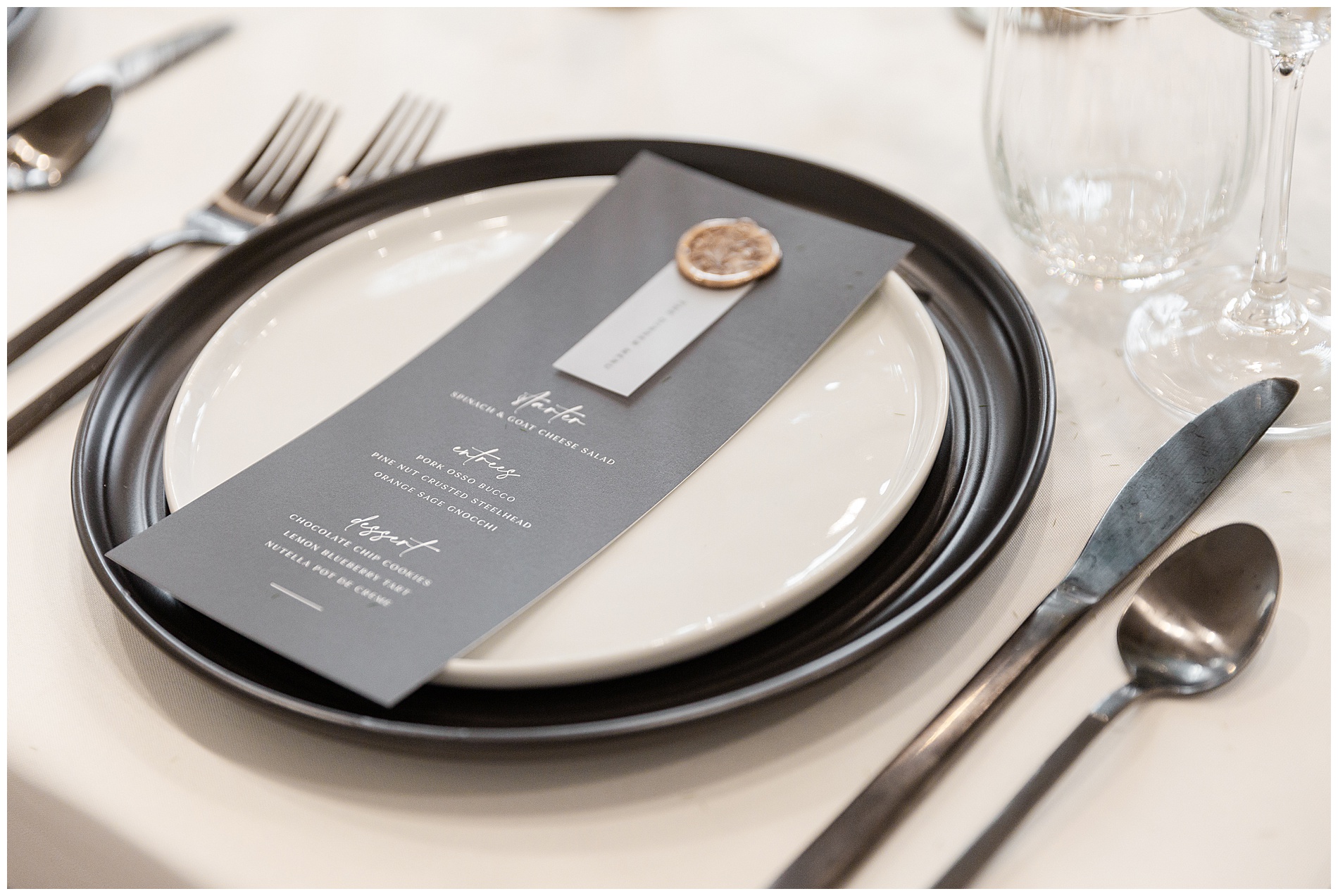 An individual place setting with black charger, white dinner plate, black or dark gray flatware, and a menu
