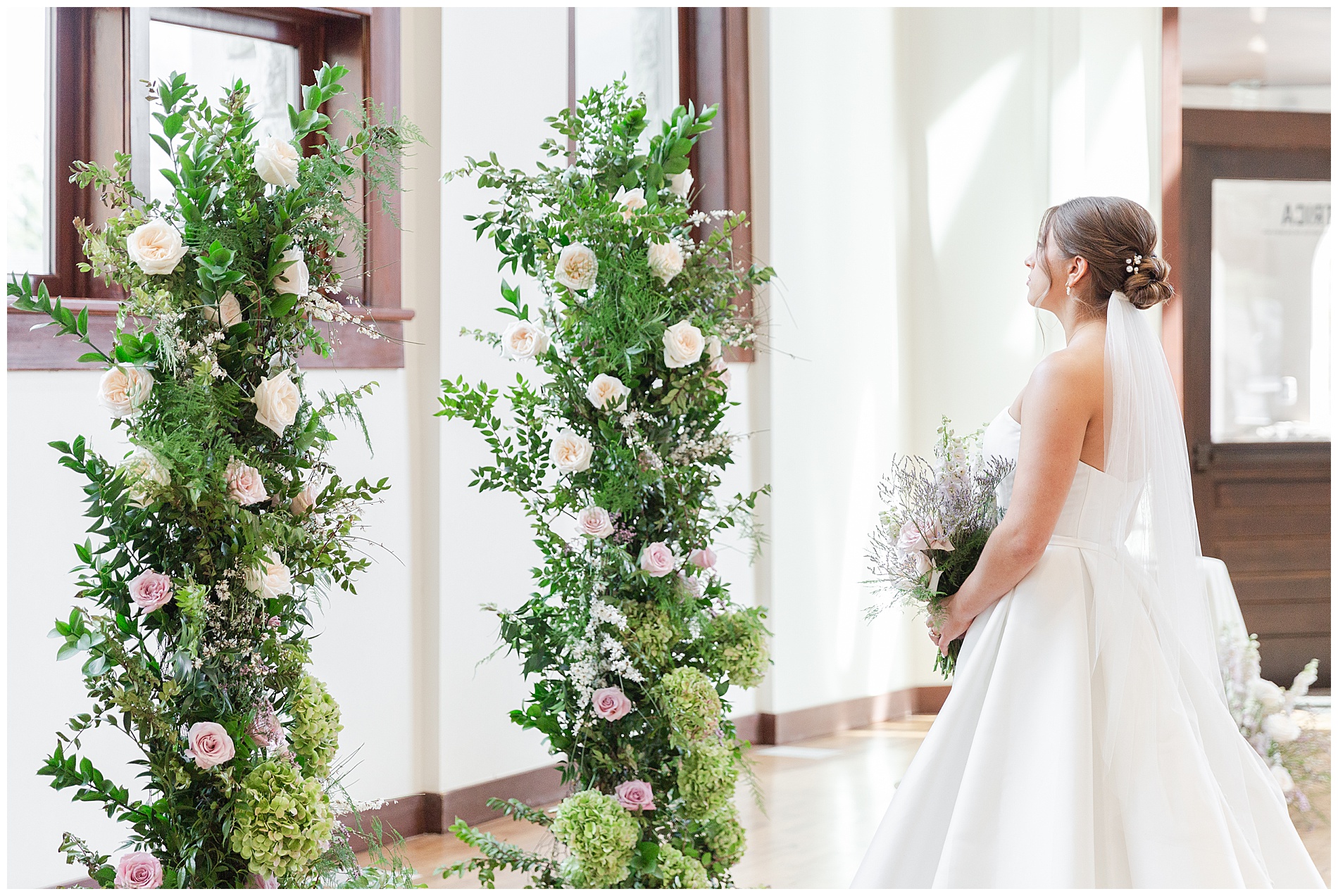 The bride stands facing the floral installations and the large window behind the ceremony area
