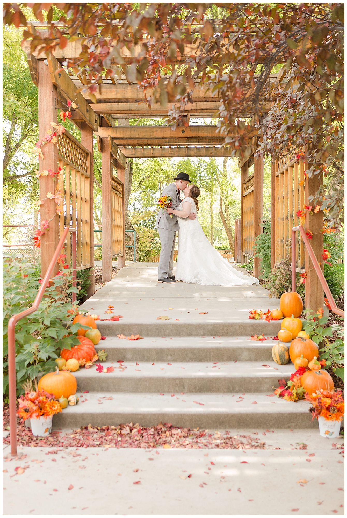 Pumpkins and fall decor line the steps to where a bride and groom kiss under a large arbor; autumn foliage in the foreground