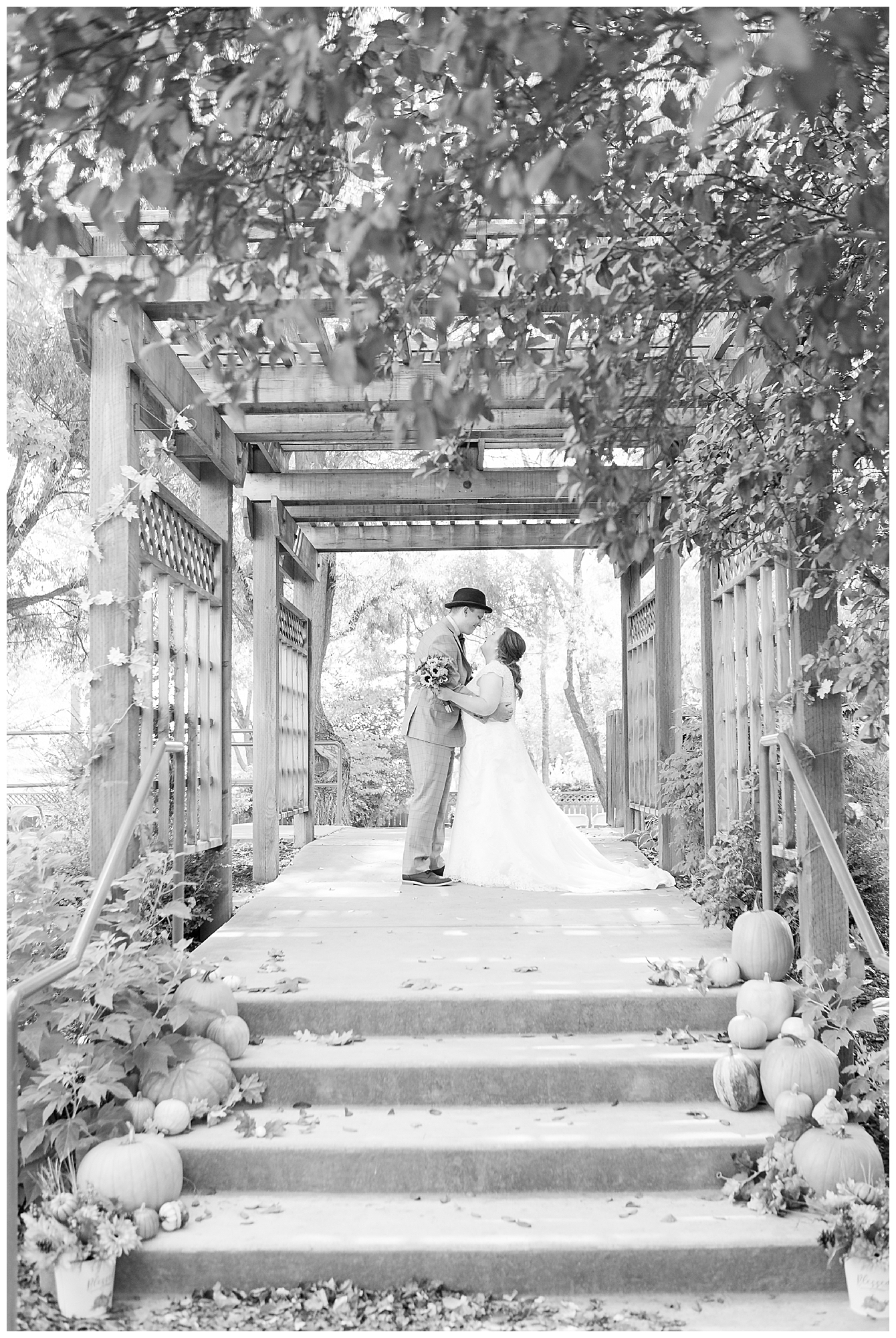 Pumpkins and fall decor line the steps to where a bride and groom stand under a large arbor, black and white