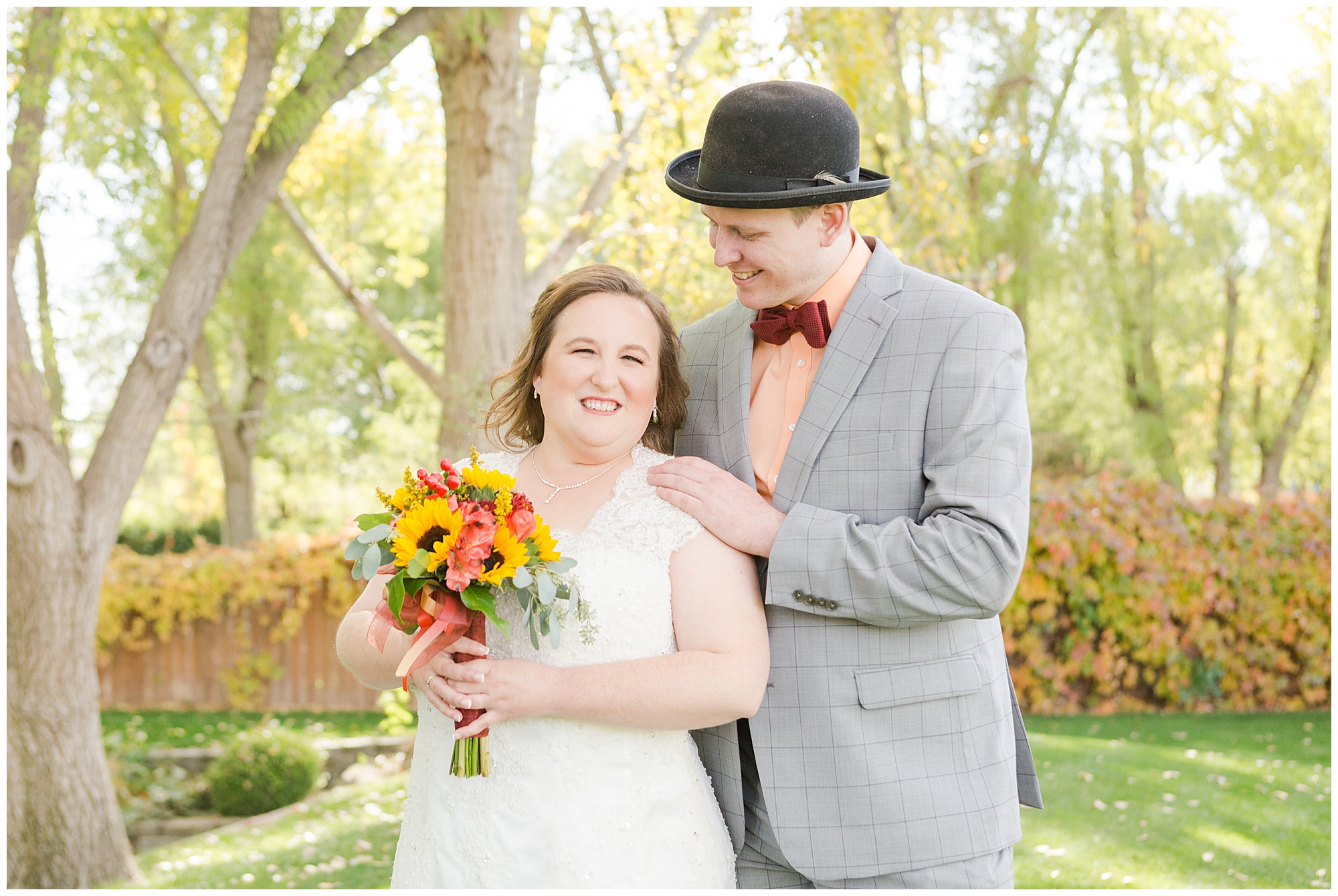 The bride smiles at the camera as her groom smiles down at her; lots of golden light filters through the autumn trees