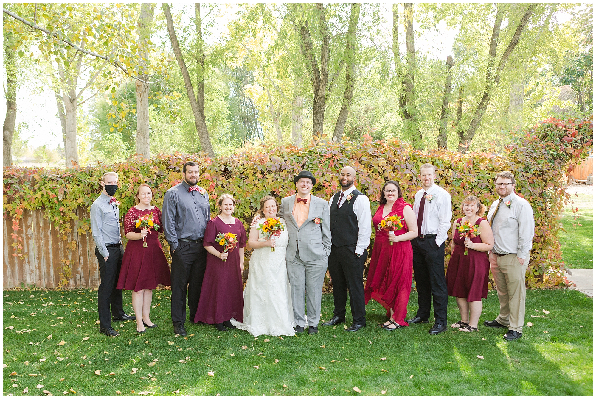 The wedding party in mismatched fall colors with a vine-covered fence and tall trees behind them