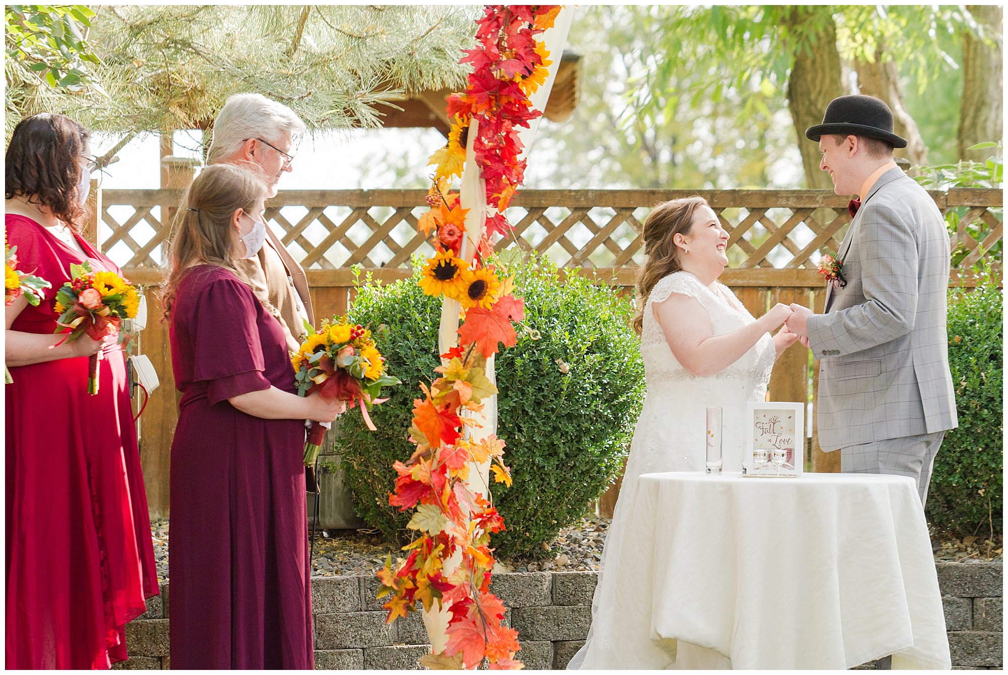 Bride and groom stand under an arch decorated with fall leaves and smile at each other during their wedding ceremony