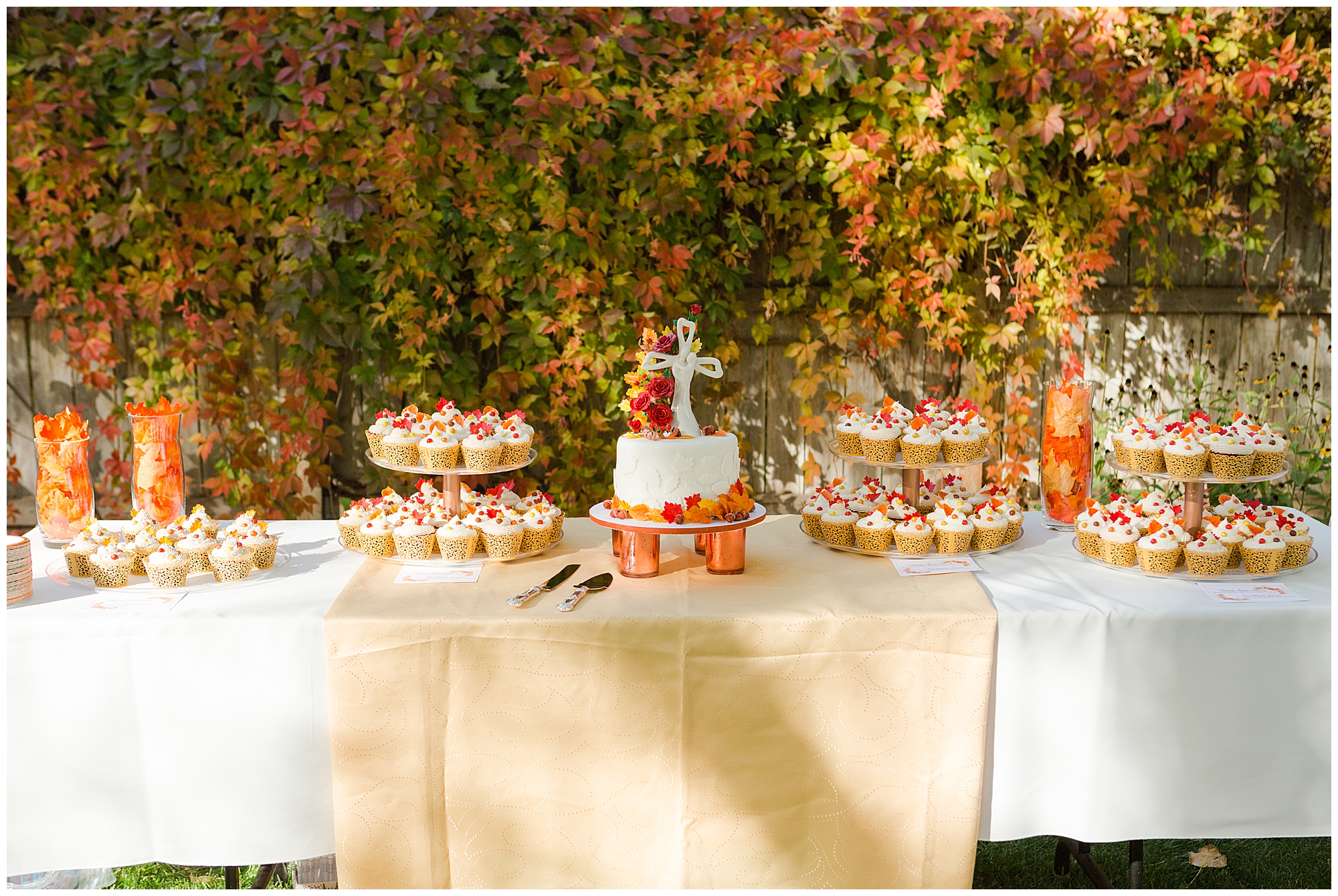 The fall-themed wedding cake and cupcakes on a display table, in front of a fence with a vine turning orange and red