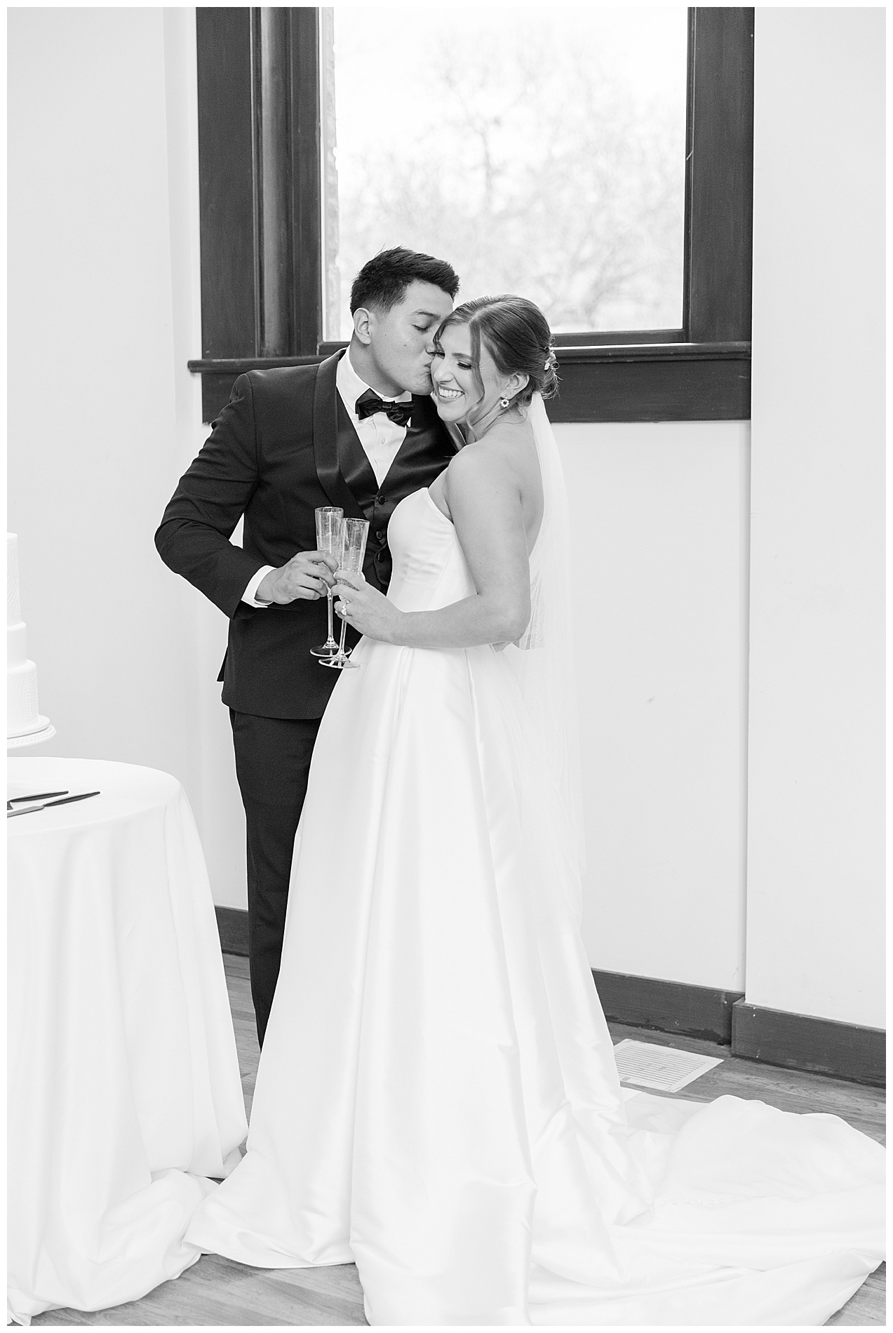 The groom kisses the bride on the cheek as they hold their champagne flutes