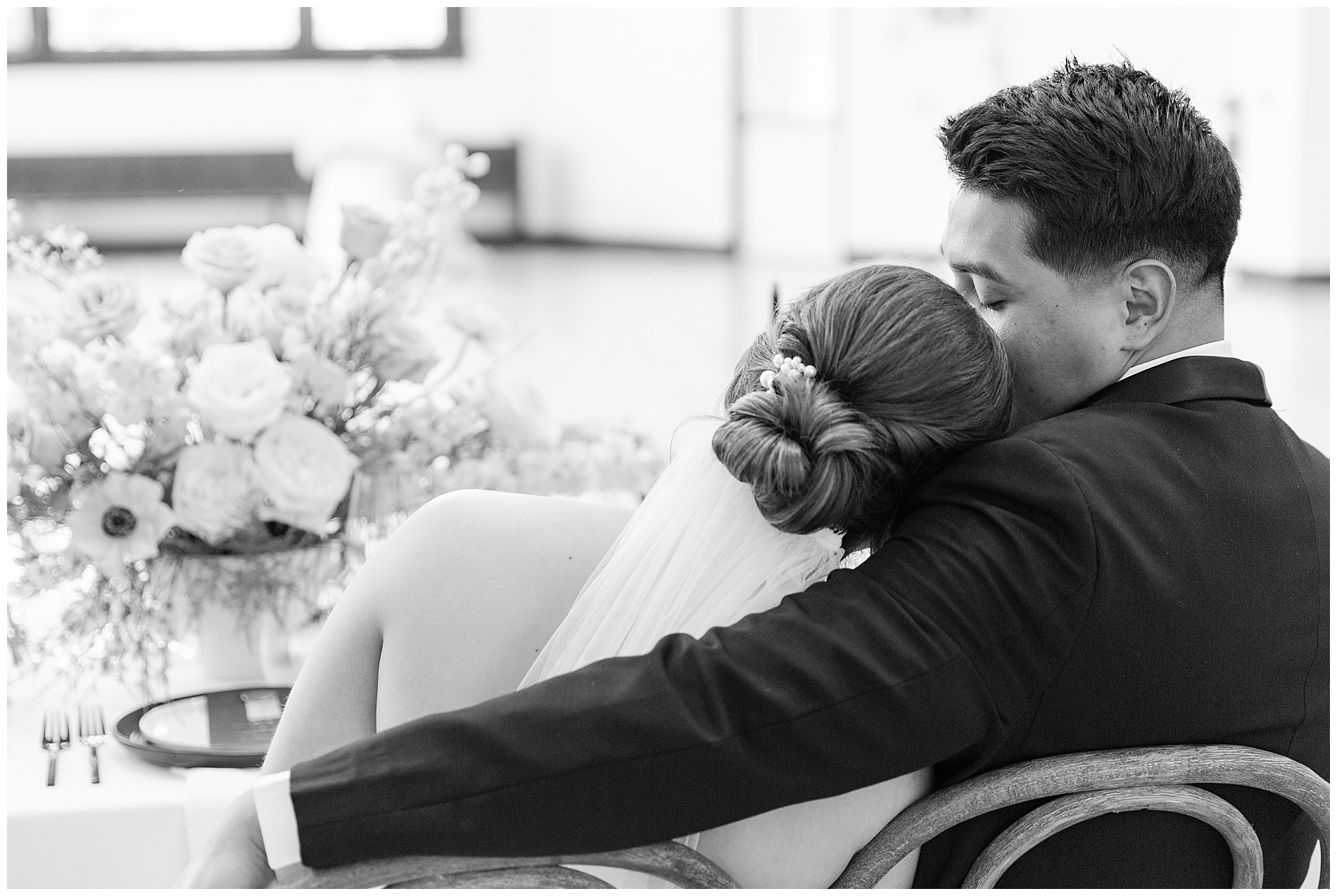 The groom puts his arm around his bride as she leans against him while they sit at their wedding reception table